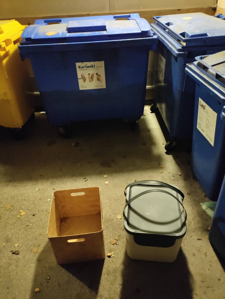 Kuva tyhjistä kierrätysastioista pahvikeräysroskiksen edestä.