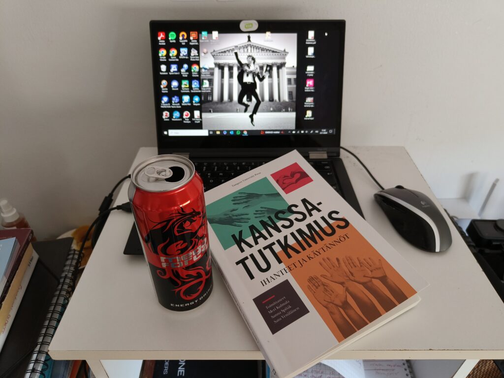 Kuva työpisteestä, jossa on tietokone, oppikirja ja energiajuomatölkki.